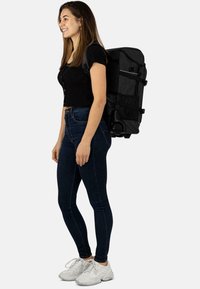 Schwarzer Rucksack aus robustem Stoff, mit verstellbaren Trägern, mehreren Fächern und Rollen, getragen von einer Person in lässiger Kleidung.