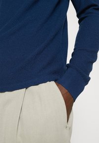 Personne portant un pull tricoté bleu à manches longues, une main dans la poche d’un pantalon beige clair sur un fond uni.