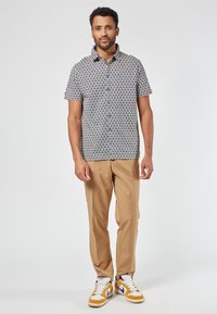 Korte mouwen knoopshirt in het blauw met een geometrisch patroon, in combinatie met beige broeken en sneakers met gele accenten.