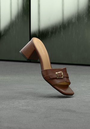 Sandale à talon en cuir marron avec bout carré, bride à boucle dorée et talon bloc sur un sol gris avec un arrière-plan vert foncé flou.