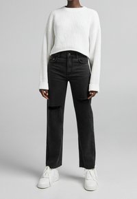 Jean noir taille haute avec genoux déchirés, associé à un pull en maille blanche texturé. Des baskets blanches complètent la tenue.