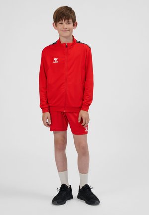 Red sports tracksuit featuring a zip-up jacket with logo, paired with shorts. Accents include black shoulder stripes and white socks. Black sneakers.