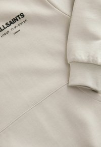 Sudadera beige con puños acanalados y cuello redondo. Presenta un logo impreso en negro y una tela texturizada con detalles de costura visibles.