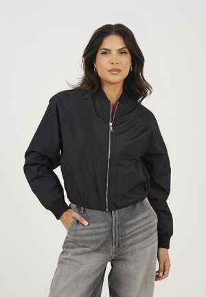 Giubbotto bomber nero con colletto alto, zip argentata e polsini a costine. Indossato con jeans grigi chiari a vita alta, caratterizzati da una vestibilità comoda.
