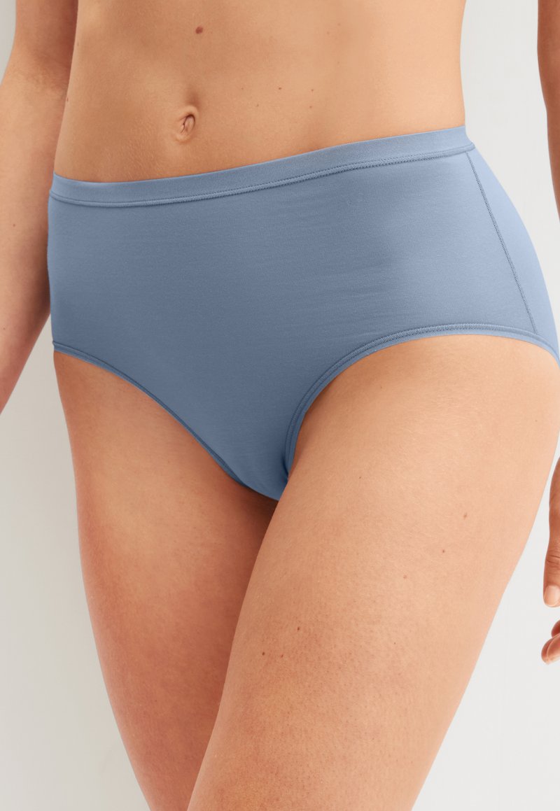 Gros plan sur une personne portant un slip taille haute en coton bleu clair, montrant le torse et la partie supérieure des cuisses sur un fond uni.