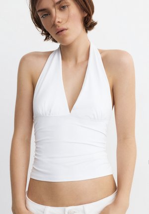 Tânără purtând un top alb sleeveless tip halter și pantaloni albi, pozând pe un fundal simplu și luminos.