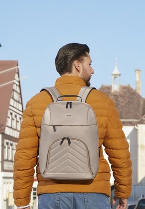 Beige backpack with a chevron-patterned front pocket and dual zippers, worn over an orange puffer jacket, background of architecture.