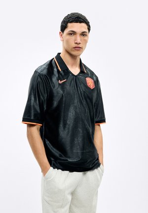 Jeune homme portant un maillot de sport noir avec des accents orange et un blason de lion, debout les mains dans les poches d'un pantalon gris clair.