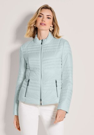 Frau trägt eine hellblaue gesteppte Jacke mit Reißverschluss und weiße Hose, posiert vor neutralem Hintergrund mit Perlenohrringen.