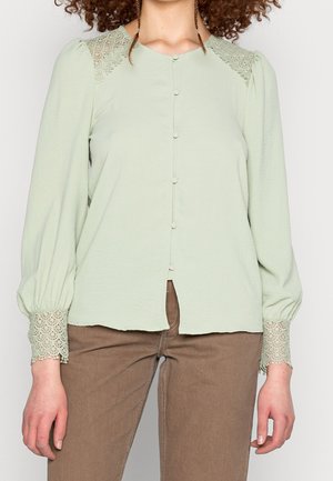 Vrouw draagt een lichtgroene blouse met lange mouwen en kanten details op de schouders en manchetten, gecombineerd met bruine broek.