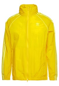 Žlutá lehká bunda s vysokým límcem, plným zipem, bílými akcenty a třemi bílými pruhy na rukávech. Má logo na hrudi.