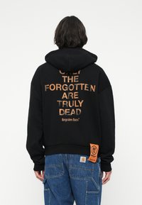 Pessoa a usar um hoodie preto com o texto laranja "SOMENTE OS ESQUECIDOS ESTÃO REALMENTE MORTOS" e etiqueta laranja, combinado com jeans de ganga azuis, de costas.