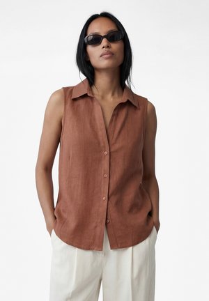 Kvinde iført ærmeløs brun knappet skjorte, hvide bukser og sorte solbriller, stående med hænderne i lommerne mod en hvid baggrund.