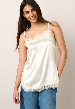 Ung kvinde med langt mørkt hår smilende, iført en cremefarvet satin camisole med blonder og blå jeans mod en lys baggrund.