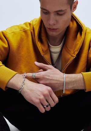 Silberschmuck umfasst ein Kettenarmband, einen strukturierten Ring und eine elegante Manschette. Das Modell trägt einen leuchtend gelben Kapuzenpullover und ein cremefarbenes Hemd.