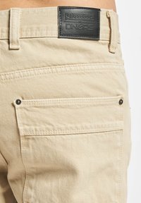 Beige denim pants featuring a black leather label, rear pockets with visible stitching, and metal hardware accents.