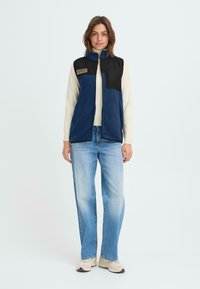 Blå fleeceställ med svarta axlar, framdragkedja, sidofickor och ett märke. Bärs över en ljuströja med blå jeans och sneakers.