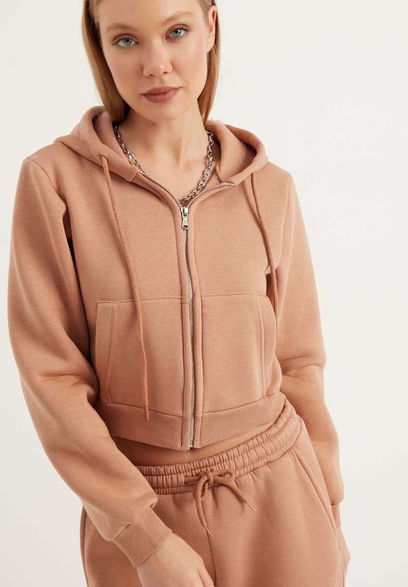 Felpa cropped con zip in morbido e leggero tessuto marrone chiaro. Presenta un cappuccio, tasche frontali, polsini a coste e una zip argentata. Abbinata a pantaloni coordinati.