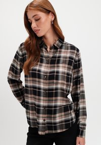 Kobieta z długimi rudymi włosami, ubrana w czarną, beżową i białą koszulę w kratę oraz czarne spodnie, patrzy w dół i na bok.