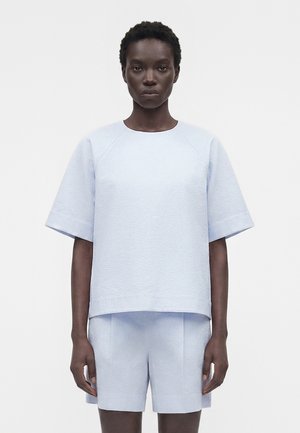 Čierna osoba oblečená v svetlomodrom textúrovanom tričku s krátkym rukávom a zodpovedajúcich kraťasoch, stojaca pred jednoduchým bielym pozadím.