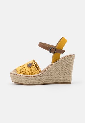 Zoccoli espadrille gialli con tacco a rocchetto, dotati di cinturino marrone alla caviglia e bottone decorativo. Presentano una piattaforma in juta testurizzata e un design con punta aperta.
