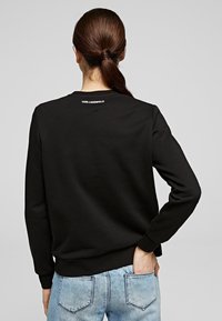 Sudadera negra con mangas largas, cuello redondo y un logo blanco en la parte trasera. Textura suave, combinada con vaqueros de mezclilla azul claro.