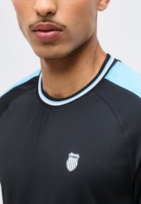 Κοντινό πλάνο ενός ατόμου που φοράει μαύρο αθλητικό πουκάμισο με ανοιχτό μπλε ρίγες στους ώμους και λογότυπο K-Swiss στο στήθος.