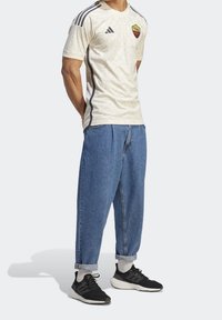 Camiseta de fútbol color crema con un patrón sutil, rayas negras de adidas y un logo del equipo rojo. Combinada con pantalones de mezclilla azules de gran tamaño y zapatillas negras.