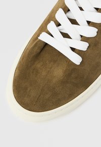 Καφέ σουέντ αθλητικό παπούτσι με λευκά κορδόνια, με υφή επιφάνειας και λαστιχένια σόλα με αντίθετη άκρη. Τακτικά ραμμένες λεπτομέρειες.