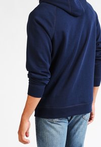 Tmavě modrá mikina s kapucí vyrobená z měkké bavlněné směsi, s žebrovanými lemy na rukávech a spodním okraji, s volným střihem a bez viditelných grafik nebo vzorů.