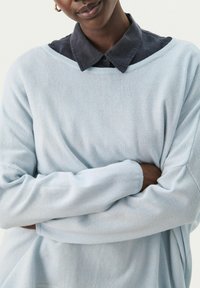 Hellblauer Pullover aus weichem Strickstoff mit weitem Rundhals und langen Ärmeln, über einem dunklen Hemd mit Kragen getragen.