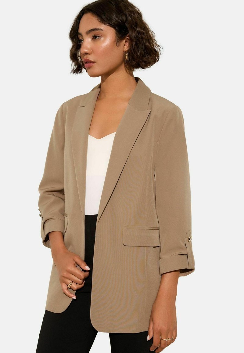 Beige sako vyrobené z lehkého materiálu, s klopovým límcem, dlouhými rukávy s knoflíky na manžetách a předními kapsami. Stylizováno přes bílé tričko.