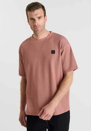 Man in een gemêleerd stof lichtroze shirt met korte mouwen en een klein zwart patch op de borst, staand tegen een effen lichte achtergrond.