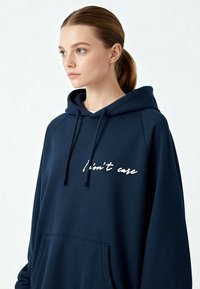 Sweat à capuche bleu marine avec une poche kangourou, arborant un texte en cursive blanc "Je m'en fiche." Conçu en tissu doux avec une coupe décontractée et une capuche ajustable.