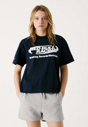 Marineblaues Baumwoll-T-Shirt mit einem weißen Grafikdesign von "RED BULL RACING" und "Breaking Records Worldwide", kombiniert mit grauen Shorts.