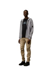Grijze zip-up hoodie over een zwart T-shirt, beige cargobroek en zwarte sneakers. Bevat een patch op de mouw en heeft een relaxte pasvorm.