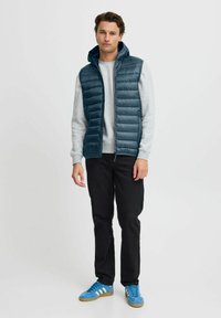 Chaleco acolchado verde oscuro con capucha sobre una sudadera gris, combinado con jeans negros y zapatillas deportivas azules con franjas blancas.