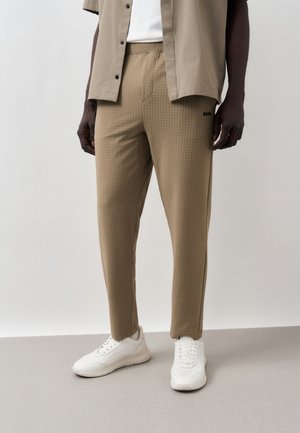 Mann trägt beige strukturierte Hose, weiße Sneakers und ein hellbeiges offenes Hemd über einem weißen Top, steht auf einem grauen Boden vor einfachem Hintergrund.