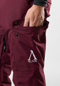 Bordowe spodnie narciarskie o gładkiej fakturze, z boczną kieszenią z zatrzaskiem oraz białym nadrukiem logo na nogawce.