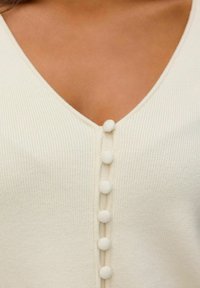 Närbild av en ribbad krämfärgad stickad topp med V-halsringning och en vertikal rad av sex tygklädda knappar.