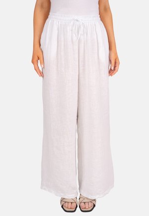 Femme portant un pantalon ample en lin blanc avec une taille élastique et un cordon de serrage, associé à des sandales beiges à bout ouvert sur un fond uni.