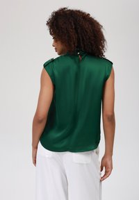 Top senza maniche in satin verde con epaulettes sulle spalle e dettaglio a chiave sulla schiena, abbinato a pantaloni bianchi, con una texture liscia e un drappeggio elegante.