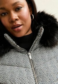Gequilted pufferjack met een zwart-wit visgraatpatroon, voorzien van een nepbont kraag en een zilveren rits. Zwarte trui eronder.