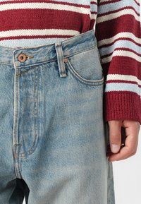Jean en denim bleu clair à taille haute, avec un bouton de fermeture et des poches avant bien visibles, associé à un pull à rayures rouges et blanches.