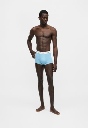 Άνδρας μοντέλο με σκούρο δέρμα, καλλίγραμμος, όρθιος ξυπόλητος, φοράει ανοιχτό μπλε εσώρουχα Calvin Klein, πάνω σε απλό ανοιχτό γκρι φόντο.