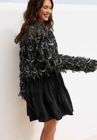 Zwarte jurk met een gelaagde, sierlijke jas met pailletten en textuurrijke, met kralen versierde accenten. De jas heeft een brede kraag en lange mouwen.