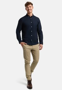 Hymyilevä mies, jolla on päällä tummansininen pikkutakki, beiget chinot ja ruskeat saappaat. Hän seisoo yhdellä kädellä taskussa yksinkertaista taustaa vasten.