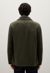 Man facing away wearing an olive green wool jacket and dark blue jeans against a plain light background.