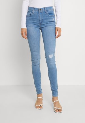 Femme portant un jean skinny bleu clair légèrement usé au niveau de la cuisse et des sandales blanches à talons hauts à brides, sur fond uni.