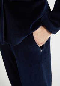 Námornícky modrá sametová dvojdielna súprava, ktorá sa skladá z priliehavého topu s vreckom a hladkou textúrou, zdôrazňujúca jemný materiál a čisté línie.